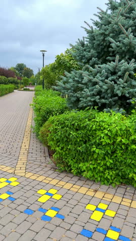 Tiled path glows with vibrant blue and yellow squares. Lush green hedges line the walkway with tall trees behind. Overcast sky casts soft light on the tranquil garden scene