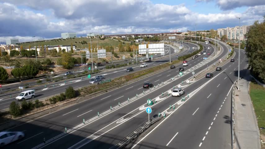 Overview of busy highway interchange featuring multiple lanes, vehicles in motion, and surrounding green landscapes under a partly cloudy sky in a developed area
