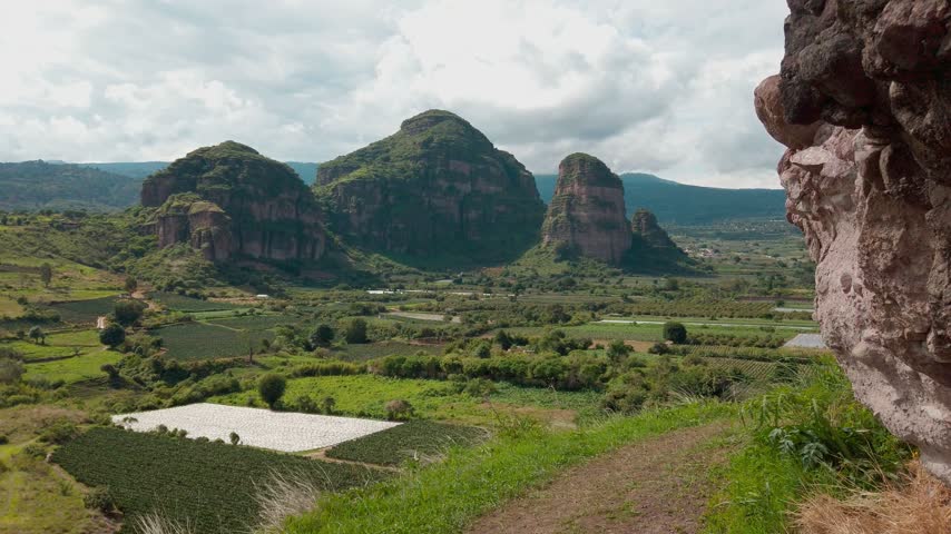 Scenic rural landscape with green farmland and distinctive rocky peaks above cultivated fields and trees beneath cloudy sky with dirt path in foreground and open land here