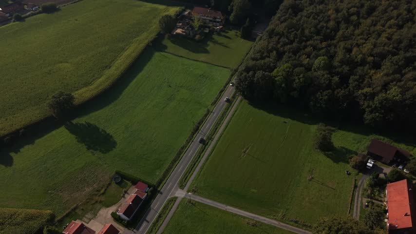 Rural road with white car driving past green meadows, dense autumn forest, farmhouse with dark roof in America, long tree shadows stretching across countryside fields. Aerial drone top down shot