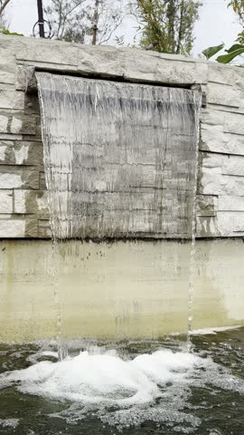 Curtain of falling water pouring down a stone-faced wall fountain into a pond, with foam at its base. Soothing footage for relaxation, meditation and landscaping projects.