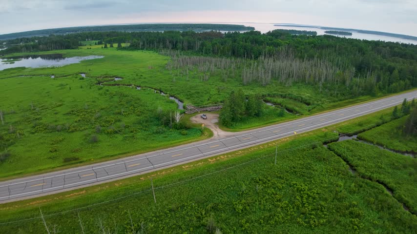 Drone view of a paved road crossing a vast green wetland with winding creeks and distant wooded Lake Huron shoreline.