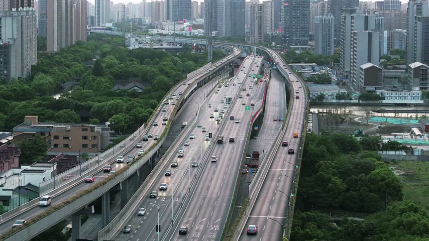 Fast-moving traffic navigates multi-lane roads surrounded by green parks and high-rise buildings in a bustling urban environment during daytime.