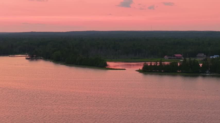 Wide aerial view of pink evening light over Lake Huron and a forested Hessel peninsula with a few shoreline homes.