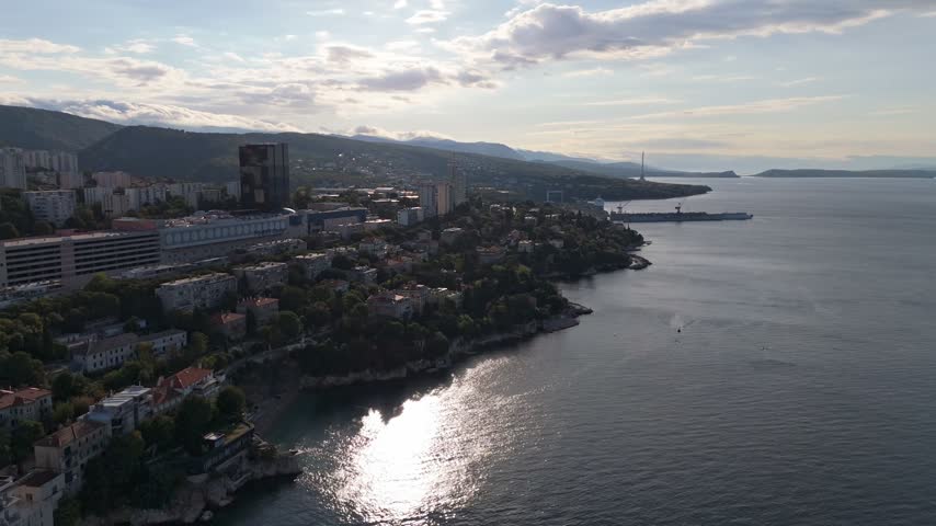 Aerial view of the coastal city of Rijeka with urban buildings, rocky shoreline, and calm sea under a soft sunset light in Rijeka, Primorje-Gorski Kotar, Croatia.