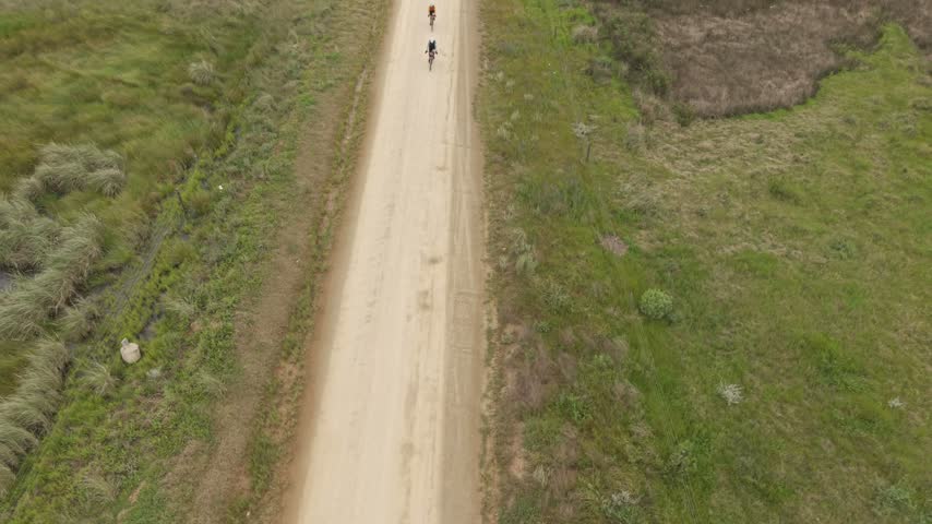 Aerial view of two cyclists riding mountain bikes along a dusty dirt road surrounded by green grassy fields in Greyton, Western Cape, South Africa.