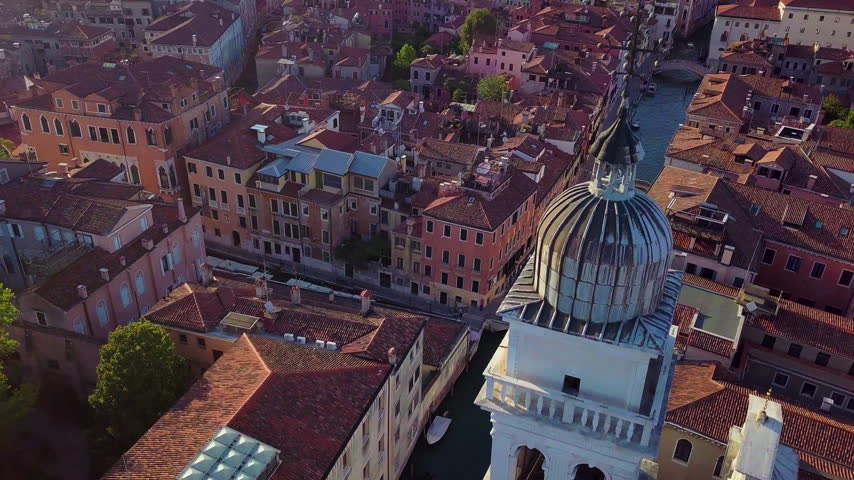 Aerial drone orbit in high-angle view around one of the twin bell towers of Santa Maria della Salute in Venice, revealing surrounding canals, rooftops and warm sun rays over the historic city.