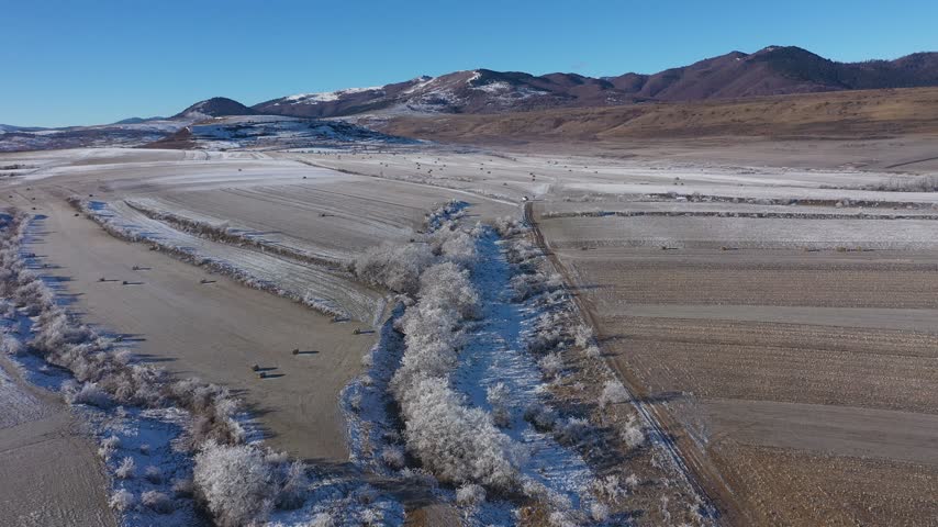   Aerial view of snow-covered field dotted with hay bales under a clear blue sky in a mountainous region during winter