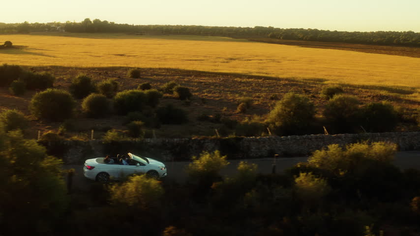 A white convertible with two passengers passes a golden wheat field in Mallorca.