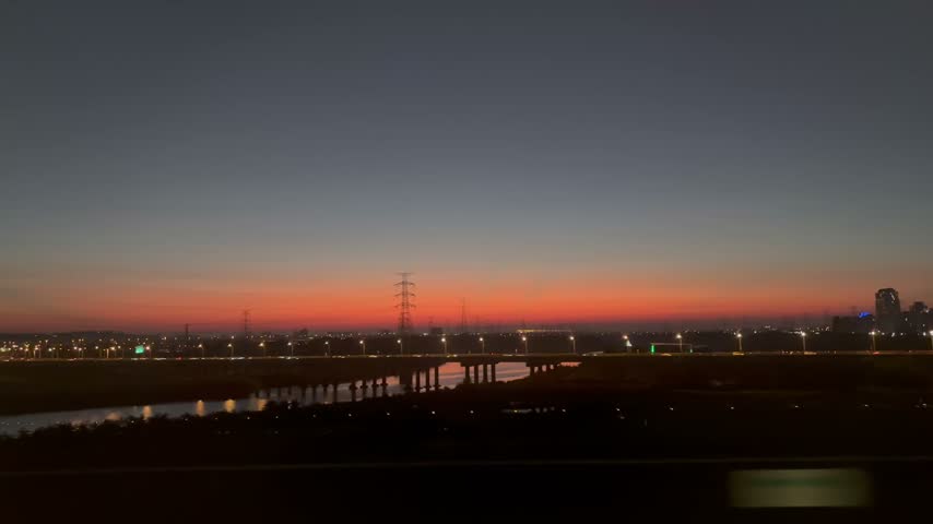 View from a high-speed train window of a vivid red and orange autumn sunset over a river in the Taichung countryside, Taiwan, with electricity pylons silhouetted against the dusk sky and city lights glowing along a bridge.