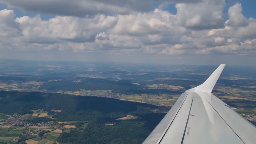 Aerial view through an airplane window as the aircraft climbs after takeoff from Zurich Airport, with the wing visible above Switzerland’s landscape, soft clouds and a distant horizon