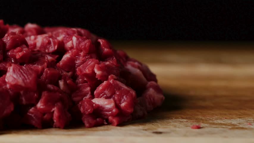 Chopped raw beef is piled on a wooden cutting board in a kitchen. The scene captures the detailed texture of the meat in preparation for a meal.