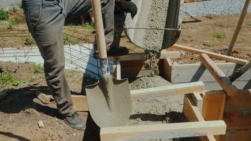 Professional construction workers pouring fresh concrete into wooden formwork to build foundation for new residential house. Industrial site activity with heavy equipment in daylight.