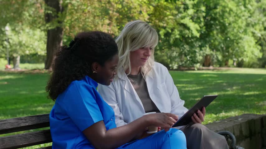 Professional female medical staff having a coffee break and discussing work on a tablet outdoors
