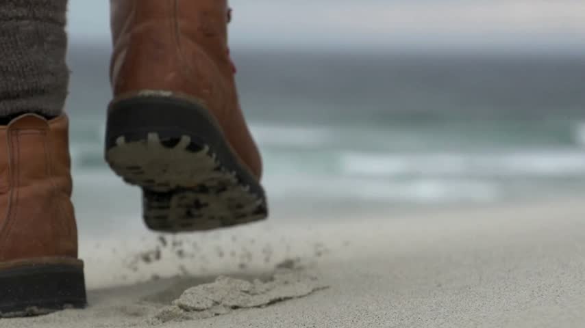 Footsteps appear in wet sand along the shore as ocean waves crash softly. A person walks slowly on the sandy beach under a cloudy sky, capturing a moment of nature.