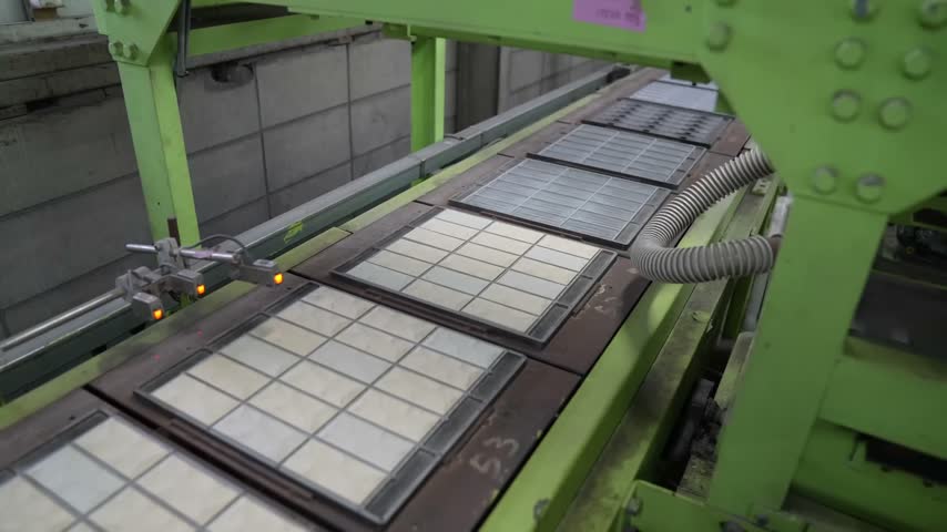 Overhead gantry arm moves above framed white tile sheets on a green ceramic production line