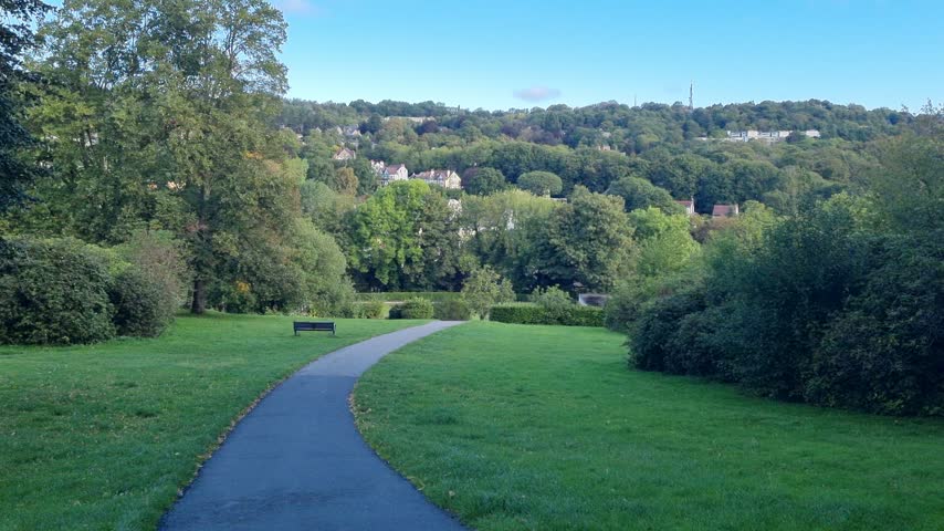 Downhill asphalt road with lawns, trees and shrubs, overlooking distant houses and a panoramic hillside landscape