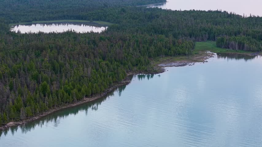 A drone glides beside a densely forested Upper Peninsula shoreline surrounding calm blue water near Hessel Michigan.