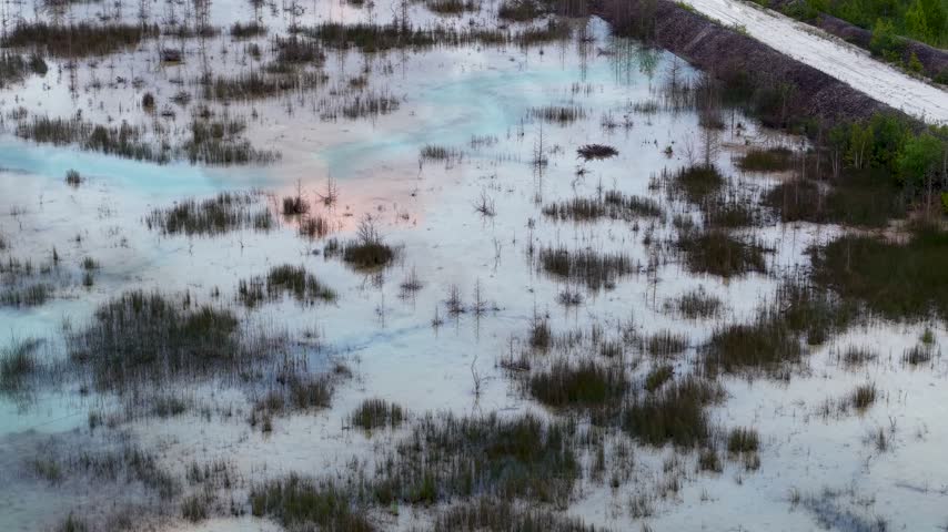 An aerial view passes over reflective shallow pools scattered through marsh vegetation beside a pale road near Hessel Michigan.