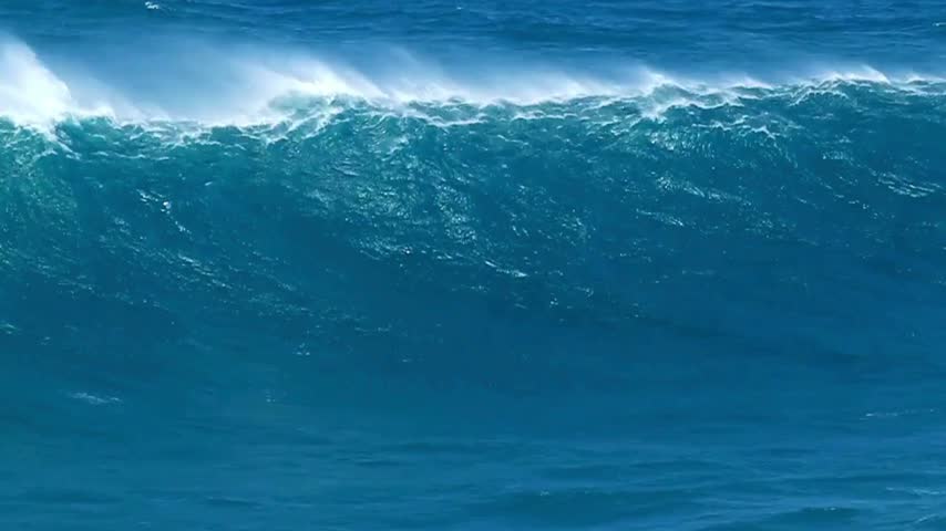 Blue ocean waves rise and fall dramatically against a rocky coastline under a clear sky. Sunlight glistens on the water surface, showcasing vivid colors of the sea.