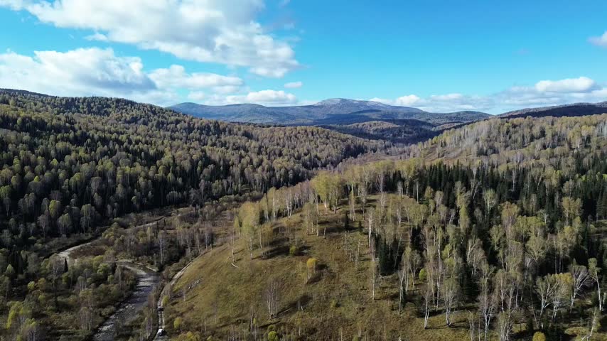 Aerial drone view of a forested mountain valley in autumn, with rolling hills covered by mixed birch and conifer forest, open grassy areas, and a winding stream running through the valley. A narrow dirt road follows the terrain, with distant mountain ridges and a blue sky with scattered clouds in the background.