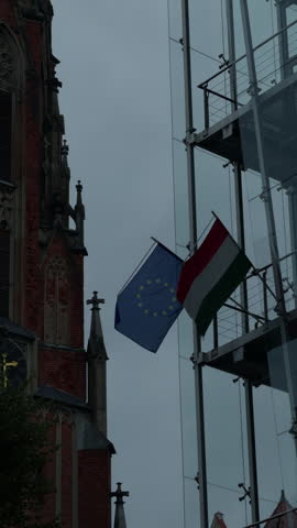 Historic brick church beside modern glass facade with European Union and Hungary flags