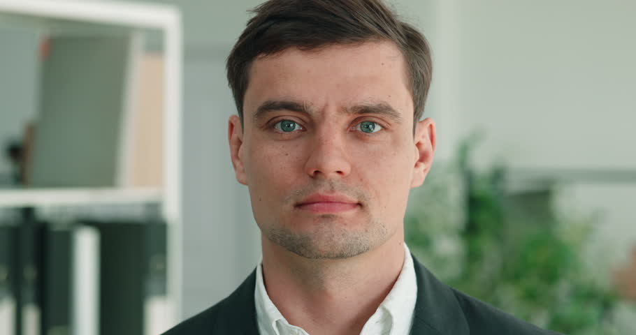 Portrait of serious Caucasian businessman in bright office keeps direct steady gaze. Man faces forward while holds composed expression during formal work moment