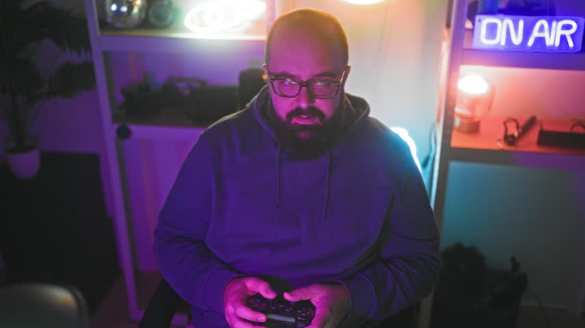 Man with beard and glasses holds a game controller with hands and makes a fist gesture while seated in a neon lit studio with an on air sign; concentration.