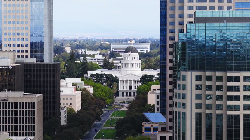 Aerial shot looking past modern downtown skyscrapers toward the historic California State Capitol dome and building in Sacramento, displaying city streets and urban trees in daytime.