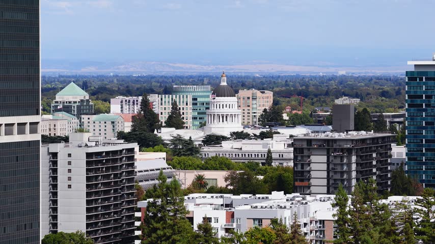 Aerial panorama of downtown Sacramento featuring the California State Capitol dome surrounded by office towers, apartment blocks, and tree canopy extending to distant horizons.