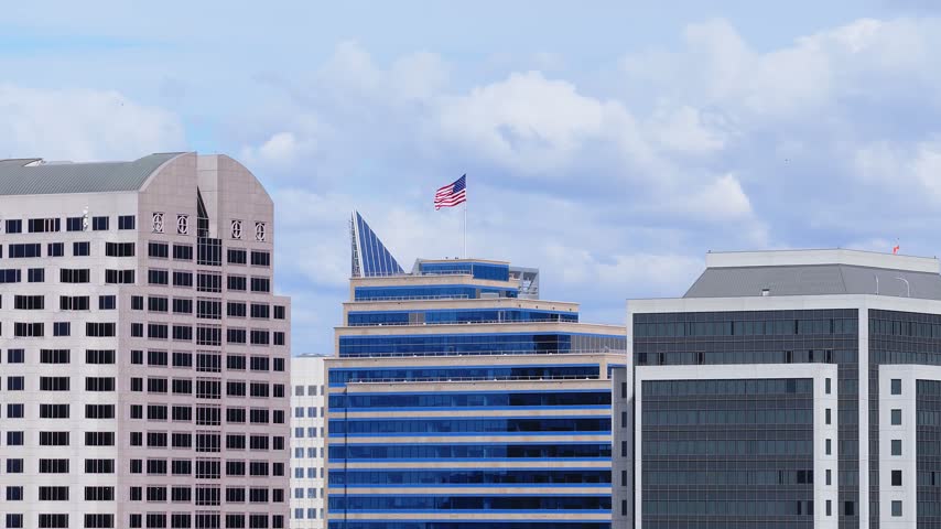 Aerial view of Sacramento downtown skyscrapers rising towards cloudy skies with an American flag fluttering atop an office building roof during daylight in California.