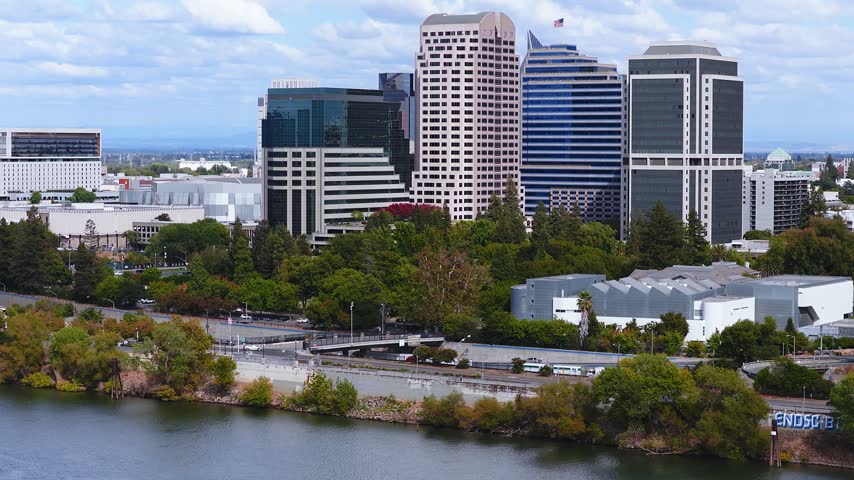 Aerial panning view of the downtown Sacramento skyline along the Sacramento River, featuring modern skyscrapers, the Crocker Art Museum, and the distant California State Capitol.