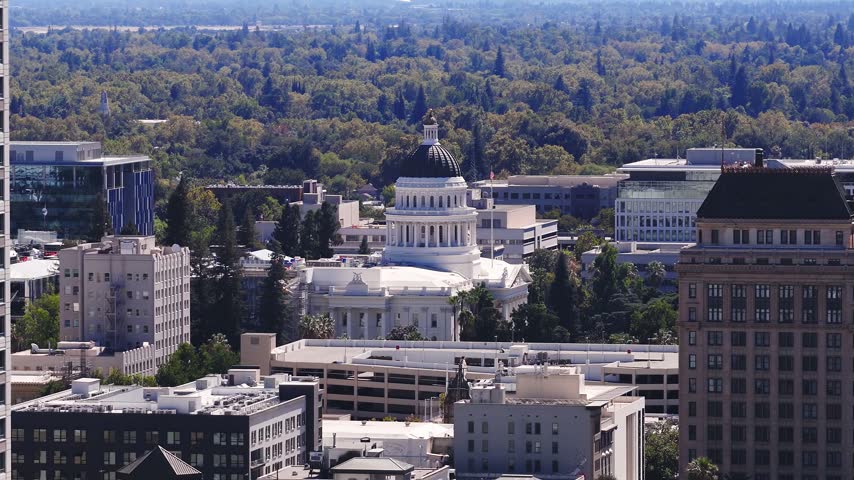Aerial view captures the neoclassical dome of the California State Capitol building rising above urban architecture and surrounding tree canopy in downtown Sacramento on a bright day.