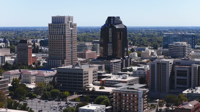 Aerial view over downtown Sacramento, California, showcasing corporate skyscrapers, office complexes, street traffic, and tree canopy stretching toward the distant horizon under clear skies.