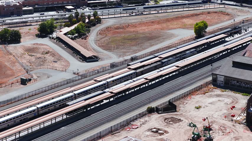 Aerial view looking down at passenger trains parked along platforms at the Sacramento Valley Station rail yard, surrounded by tracks, maintenance areas, and industrial structures in California.