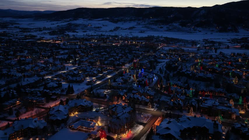 Blue hour drone view of a winter resort resembling Breckenridge, Colorado, captures snow covered homes, festive trees, warm streetlights, and dark Rocky Mountain ridges after a snowfall.