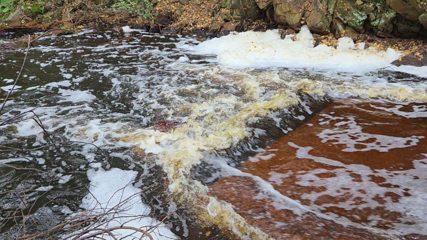 Close-up of river rapids below an old dam in the Urals: dark peat water breaks over a reddish stone ledge into swirling white foam. Natural water texture background.