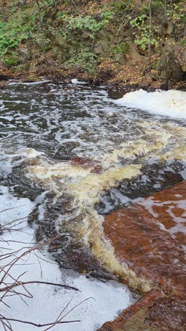 Vertical close-up of a turbulent river step below the Kyshtym dam: brown peat water surges over rocks, throwing up white foam, with a mossy stone wall and shrubs on the bank.