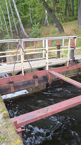 Old dam of the Kyshtym pond, Ural: a wooden sluice gate with a chain lifting mechanism, red steel beams, a white footbridge and birch forest, dark water flowing below. Vertical video.