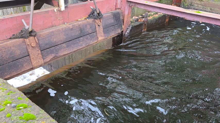 Top view of the sluice of a historic dam in Kyshtym, Ural: dark water streams past a rusty wooden gate with red steel supports, foam collecting at the mossy concrete edge.
