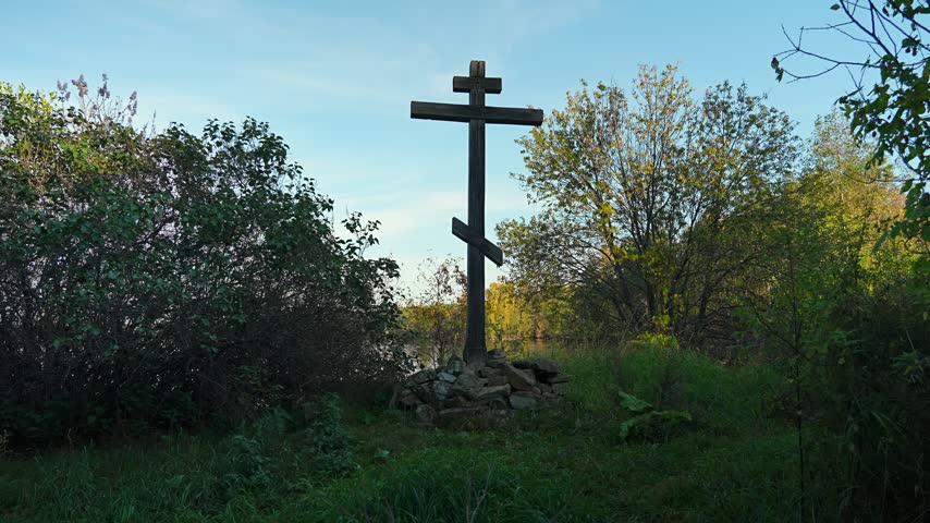 A large wooden Christian cross on the shore of a lake, surrounded by greenery, against a backdrop of a cloudy blue sky. Religious concept.