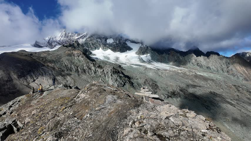 View of Grossglockner from near Studl Hut in the Austrian Alps, dramatic glaciated mountain landscape with hikers on a rocky ridge beneath clouds and blue sky