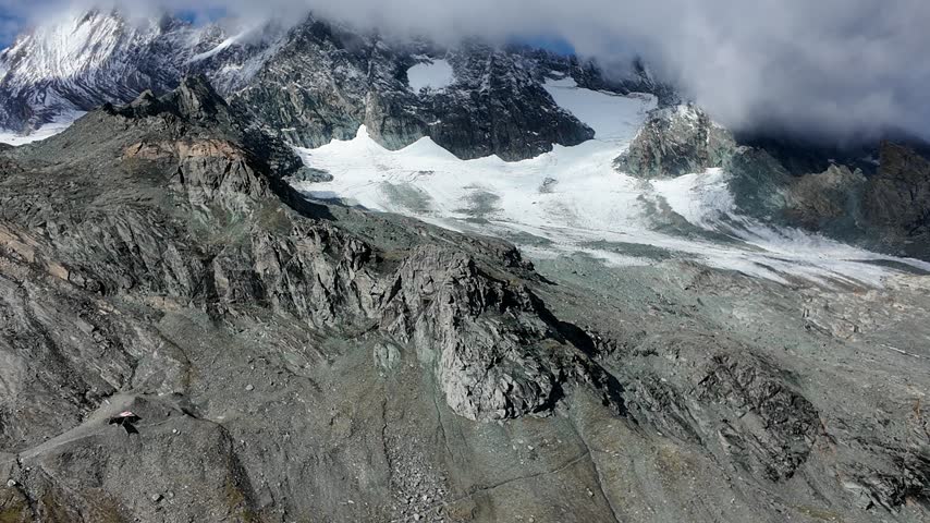 View of Grossglockner from near Studl Hut in the Austrian Alps, dramatic glaciated mountain landscape with hikers on a rocky ridge beneath clouds and blue sky