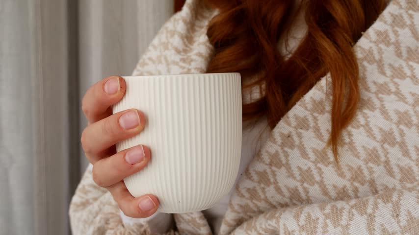 Woman holding hot drink while wrapped in warm blanket for cozy relaxation at home.