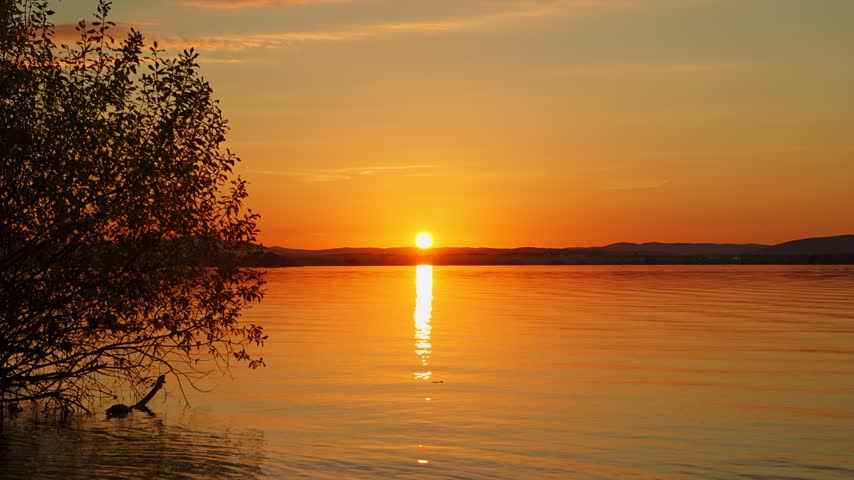 A beautiful sunset landscape with a view of the lake during the golden hour. A mirror‑like reflection of the sunset’s colors on the water. A unique natural beauty.