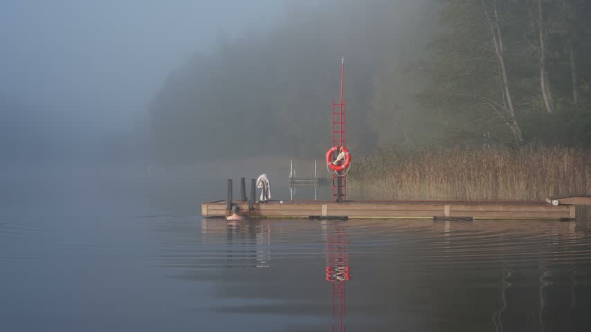 Middle aged man entering cold lake water from wooden dock early autumn morning foggy landscape Finland