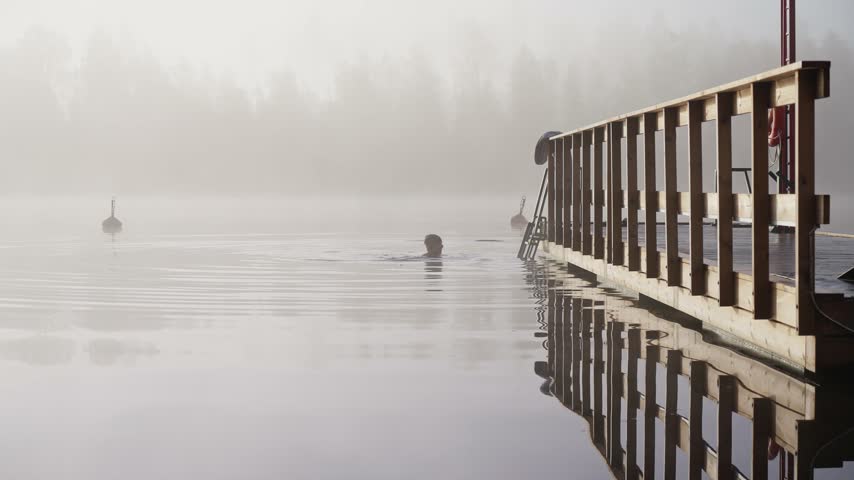 Middle aged man entering cold lake water from wooden dock early autumn morning foggy landscape Finland