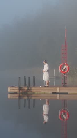 Middle aged man entering cold lake water from wooden dock early autumn morning foggy landscape Finland