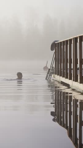Middle aged man entering cold lake water from wooden dock early autumn morning foggy landscape Finland