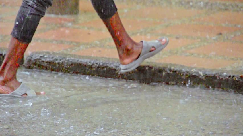 Close up view of bare feet wearing casual sandals walking through flooded tiled ground during tropical rain shower with water splashing against legs while person moves along street sidewalk